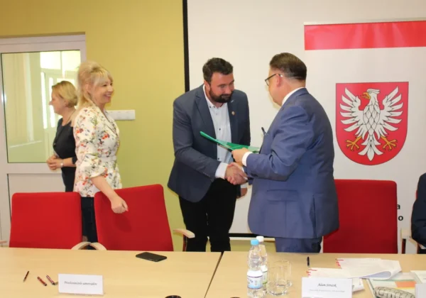 Dwie osoby w garniturach ściskają sobie dłonie podczas spotkania w auli, obok uśmiechnięta kobieta, a w tle widoczny jest baner z herbem i czerwone krzesła – scena związana z dofinansowaniem dróg dojazdowych do gruntów rolnych.