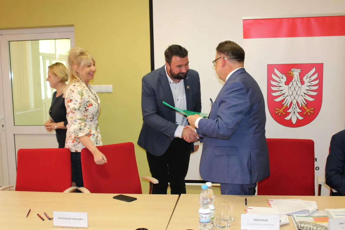 Dwie osoby w garniturach ściskają sobie dłonie podczas spotkania w auli, obok uśmiechnięta kobieta, a w tle widoczny jest baner z herbem i czerwone krzesła – scena związana z dofinansowaniem dróg dojazdowych do gruntów rolnych.