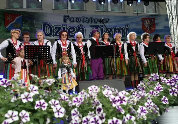 Zespół ludowy w tradycyjnych, kolorowych strojach stoi na scenie podczas jubileuszowego Powiatowego Dnia Ziemi w Słubicach, z kwiatami w pierwszym planie.