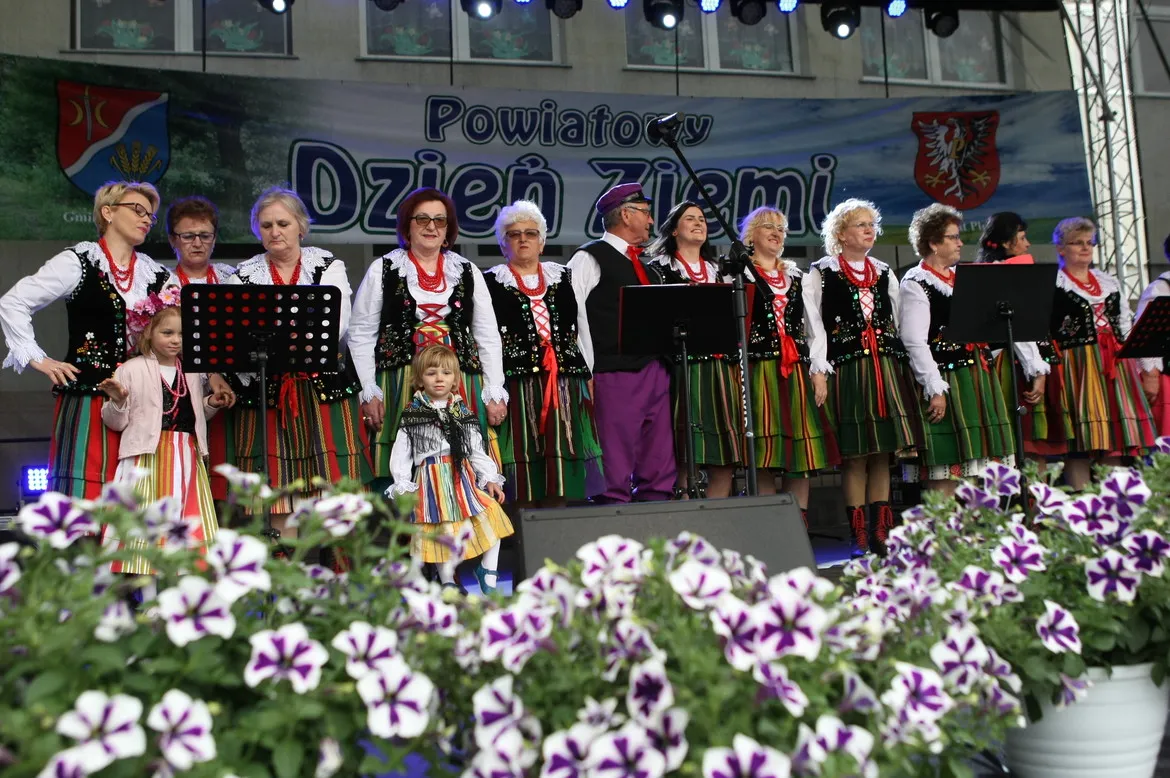 Zespół ludowy w tradycyjnych, kolorowych strojach stoi na scenie podczas jubileuszowego Powiatowego Dnia Ziemi w Słubicach, z kwiatami w pierwszym planie.