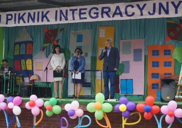 Na scenie podczas XIII edycji Letniego Pikniku Integracyjnego w Goślicach trzy osoby mówią do mikrofonów, a w tle widać kolorową dekorację w postaci budynków i balony na scenie.