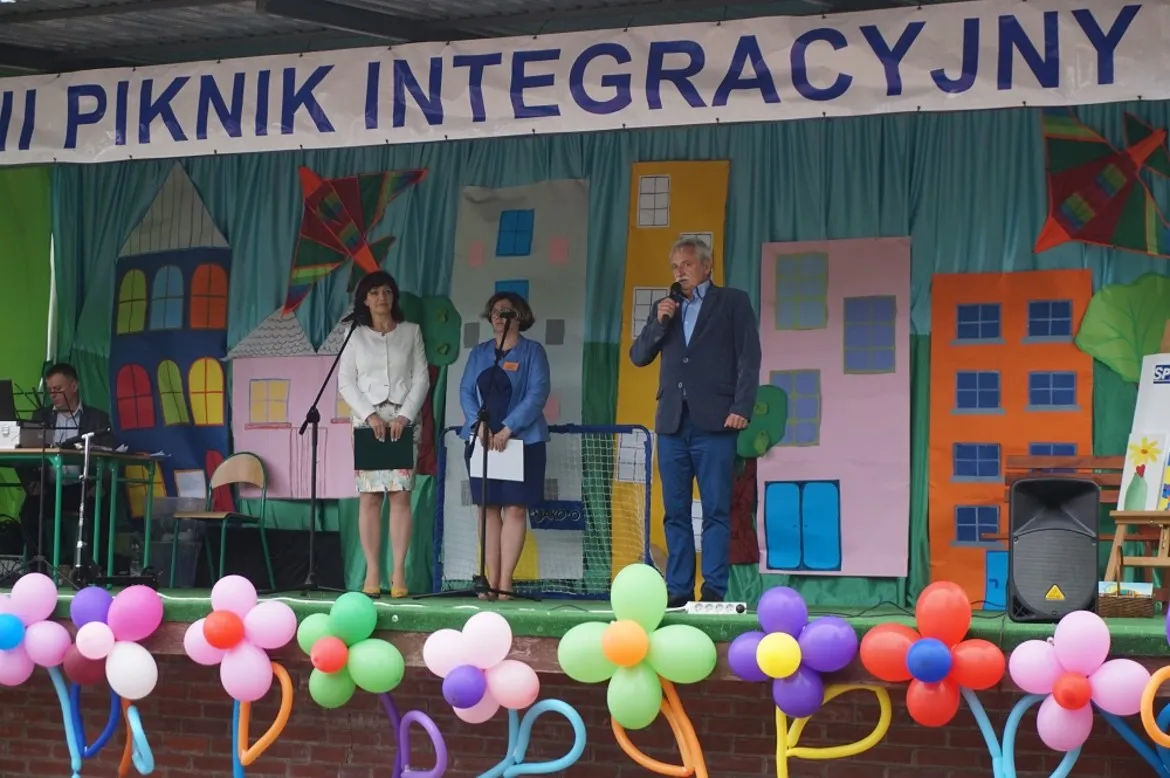 Na scenie podczas XIII edycji Letniego Pikniku Integracyjnego w Goślicach trzy osoby mówią do mikrofonów, a w tle widać kolorową dekorację w postaci budynków i balony na scenie.