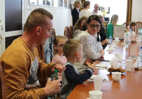 Grupa dorosłych i dzieci siedzi przy długim stole podczas uroczystości rozstrzygnięcia XI edycji Powiatowego Konkursu Fotograficznego, z napojami i przekąskami na stole.