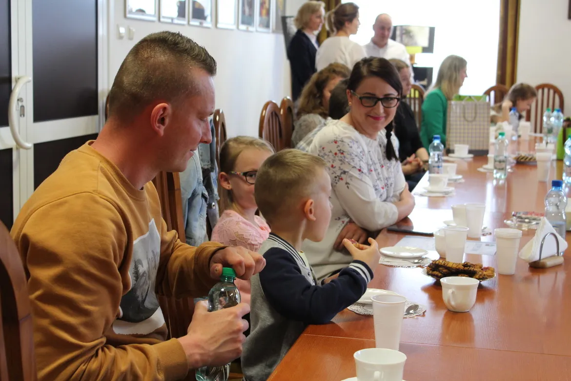 Grupa dorosłych i dzieci siedzi przy długim stole podczas uroczystości rozstrzygnięcia XI edycji Powiatowego Konkursu Fotograficznego, z napojami i przekąskami na stole.