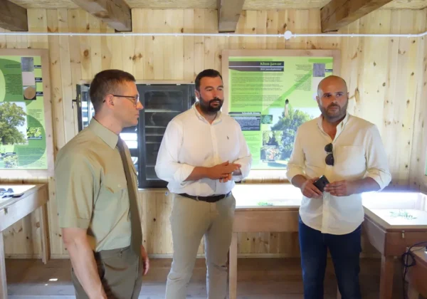 Trzech mężczyzn w jasnych koszulach stoi w drewnianej sali wystawowej z zielonymi plakatami informacyjnymi w tle, podczas Dnia w gminie Łąck.