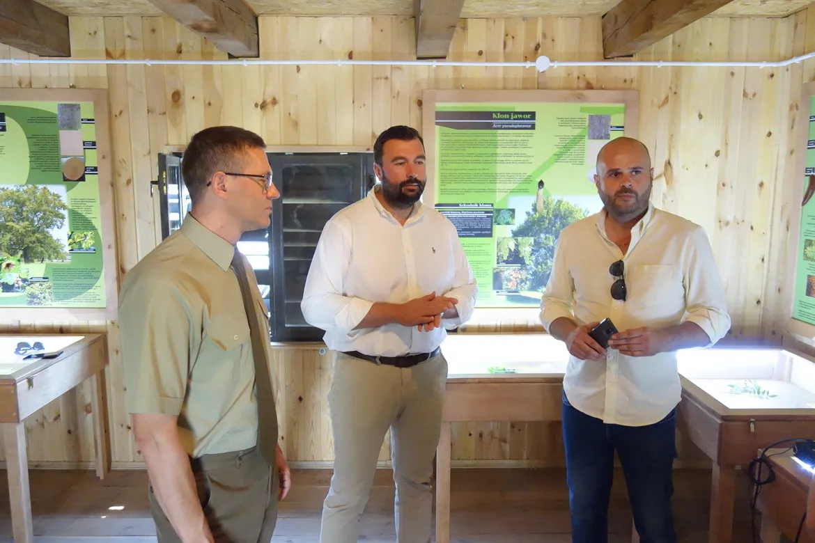 Trzech mężczyzn w jasnych koszulach stoi w drewnianej sali wystawowej z zielonymi plakatami informacyjnymi w tle, podczas Dnia w gminie Łąck.