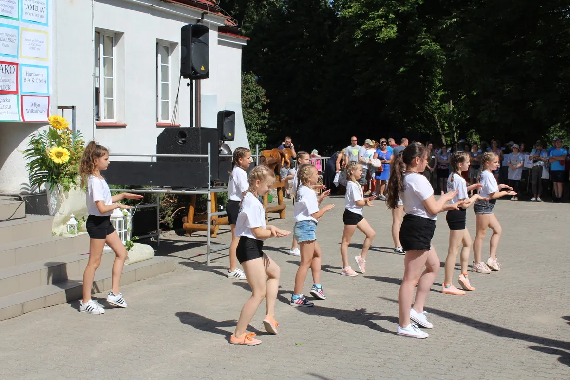 Grupa dziewcząt w białych koszulkach i czarnych szortach tańczy na placu przed sceną podczas XXVII Letniej Olimpiady Sportowej Osób Niepełnosprawnych w Nowym Miszewie, przed zgromadzoną publicznością.