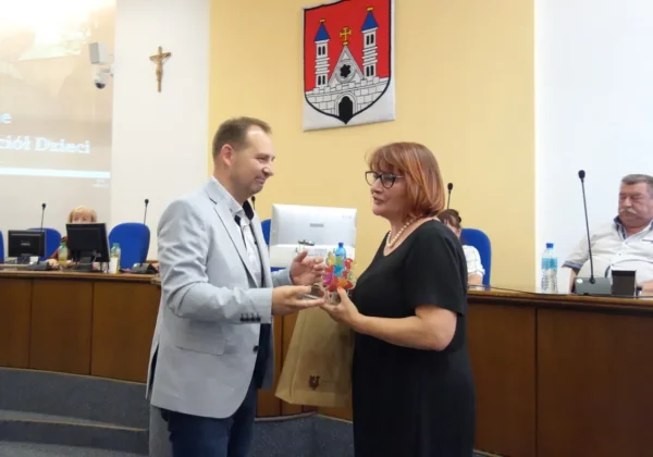 Uroczystość z okazji 100-lecia Towarzystwa Przyjaciół Dzieci: mężczyzna w jasnoszarym garniturze wręcza kobiecie kolorowy upominek, stojąc przy stole konferencyjnym, z herbem miasta widocznym na tle.