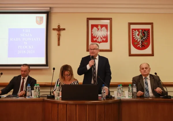 Uczestnicy VIII sesji Rady Powiatu w Płocku: mężczyzna w garniturze przemawia do mikrofonu stojąc przy stole, obok niego siedzą dwie osoby; w tle wyświetla się prezentacja z napisem „VIII Sesja Rady Powiatu w Płocku 2018-2023”, a na ścianie wiszą krzyż i dwa herby w ramkach.