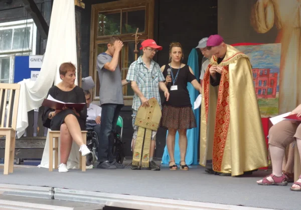 Na scenie podczas uroczystości patronalnych w DPS Brwilno grupa osób, w tym ksiądz w złotym ornacie, błogosławi zgromadzonych, podczas gdy inni odczytują materiały i prowadzą wystąpienia.