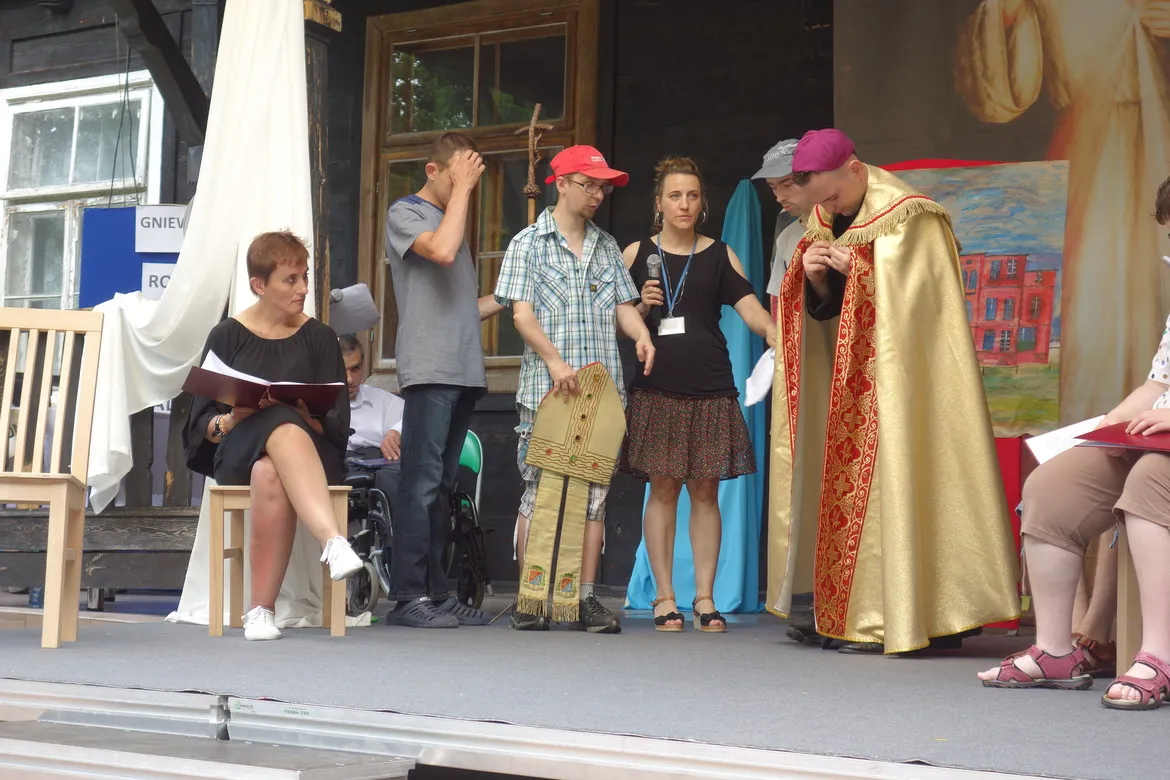 Na scenie podczas uroczystości patronalnych w DPS Brwilno grupa osób, w tym ksiądz w złotym ornacie, błogosławi zgromadzonych, podczas gdy inni odczytują materiały i prowadzą wystąpienia.