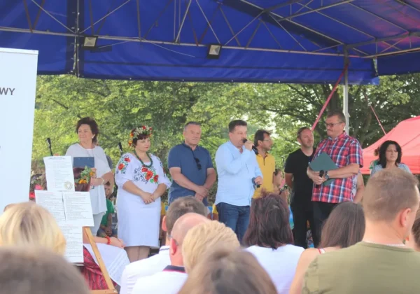 Grupa ludzi na scenie pod niebieskim namiotem podczas Biesiady Świętojańskiej w Nowym Duninowie, wśród nich prowadzący z mikrofonem i kobieta w tradycyjnym stroju ludowym, a widownia w pierwszym planie.