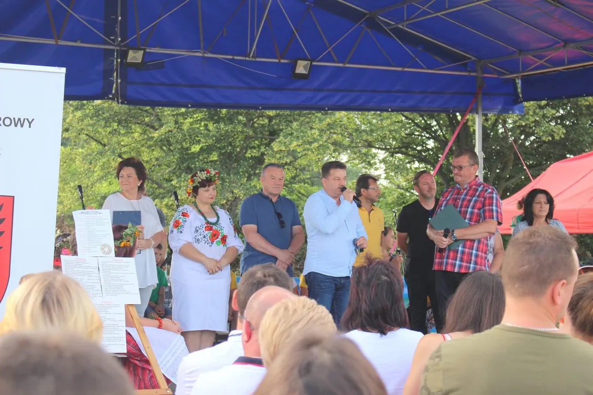 Grupa ludzi na scenie pod niebieskim namiotem podczas Biesiady Świętojańskiej w Nowym Duninowie, wśród nich prowadzący z mikrofonem i kobieta w tradycyjnym stroju ludowym, a widownia w pierwszym planie.