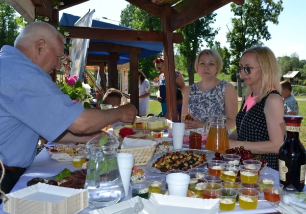 Mężczyzna w niebieskiej koszuli serwuje jedzenie i napoje przy stoisku pełnym ciast, napojów i owoców, podczas XV Jarmarku Norbertańskiego, obok stoją dwie kobiety.