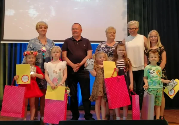 Na scenie podczas Dnia radości i śpiewu w Radzanowie grupa dorosłych i dzieci odbiera nagrody i kolorowe torby prezentowe.