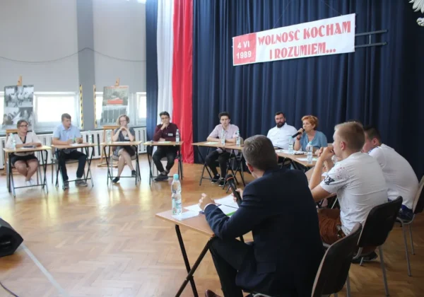 Panel dyskusyjny w dużej sali: uczestnicy siedzą przy stołach ustawionych w półkole, w tle baner z napisem „Wolność kocham i rozumiem…” i datą 4 VI 1989.