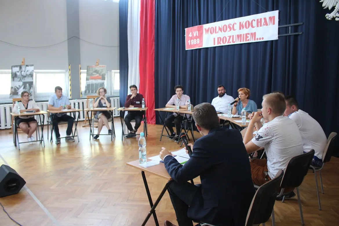 Panel dyskusyjny w dużej sali: uczestnicy siedzą przy stołach ustawionych w półkole, w tle baner z napisem „Wolność kocham i rozumiem…” i datą 4 VI 1989.