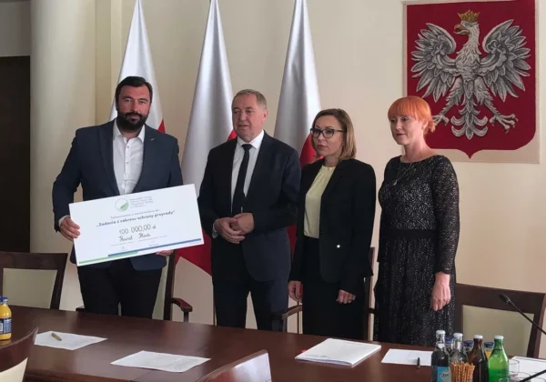 Czterech przedstawicieli samorządowych stoi przy stole podczas podpisywania umowy o dofinansowanie zabiegów pielęgnacyjnych drzewostanu i nasadzeń w DPS Koszelew oraz w Brwilnie, trzymając duży czek na 100 000 zł.