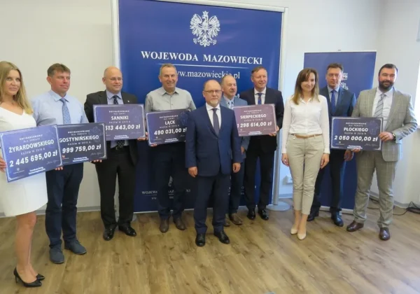 Grupa urzędników i samorządowców stoi w sali, trzymając plakietki z kwotami dofinansowania Funduszu Dróg Samorządowych, na tle baneru z napisem Wojewoda Mazowiecki, ogłaszającego ponad 9,7 mln zł dla subregionu płockiego.