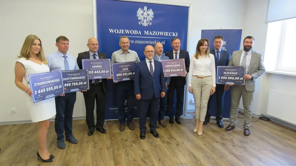 Grupa urzędników i samorządowców stoi w sali, trzymając plakietki z kwotami dofinansowania Funduszu Dróg Samorządowych, na tle baneru z napisem Wojewoda Mazowiecki, ogłaszającego ponad 9,7 mln zł dla subregionu płockiego.
