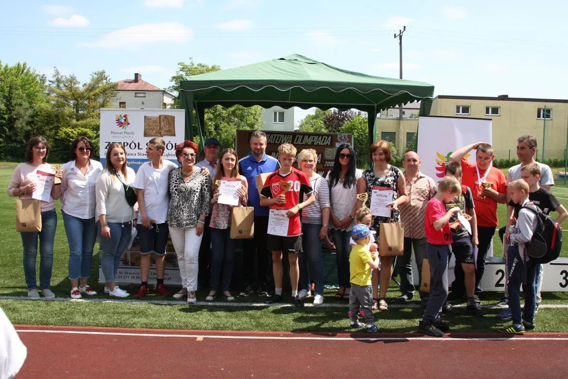 Grupa uczestników i organizatorów stoi na boisku obok zielonego namiotu podczas IX Powiatowej Olimpiady Integracyjnej 2019 w Gąbinie, wręczając dyplomy i upominki.