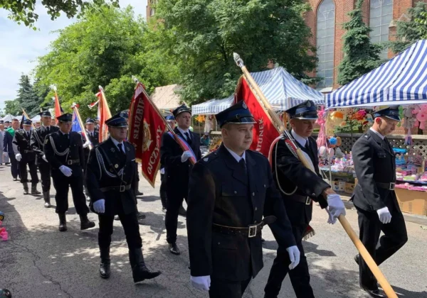 Grupa mężczyzn w czarnych mundurach i białych rękawiczkach maszeruje w paradzie z flagami ulicą podczas XX Dni Bielska, w tle stragany i namioty w paski.