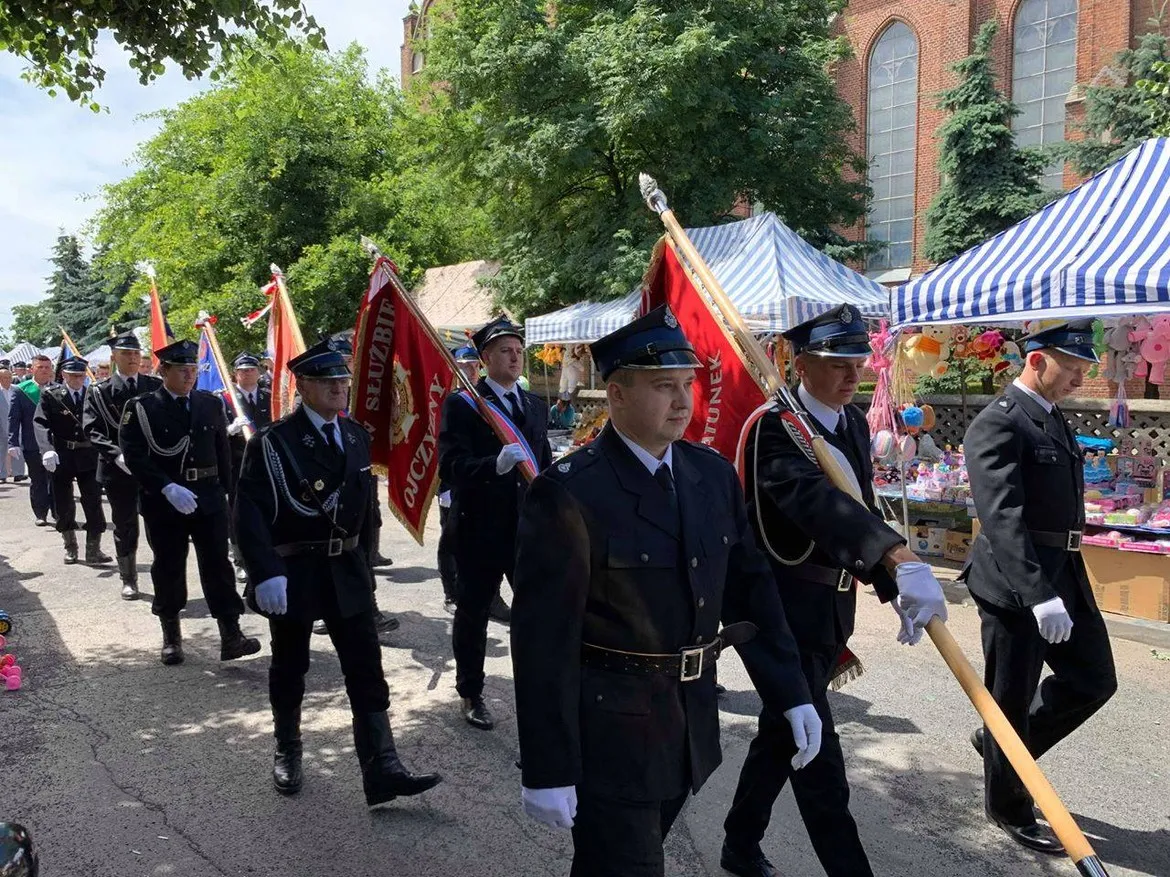 Grupa mężczyzn w czarnych mundurach i białych rękawiczkach maszeruje w paradzie z flagami ulicą podczas XX Dni Bielska, w tle stragany i namioty w paski.