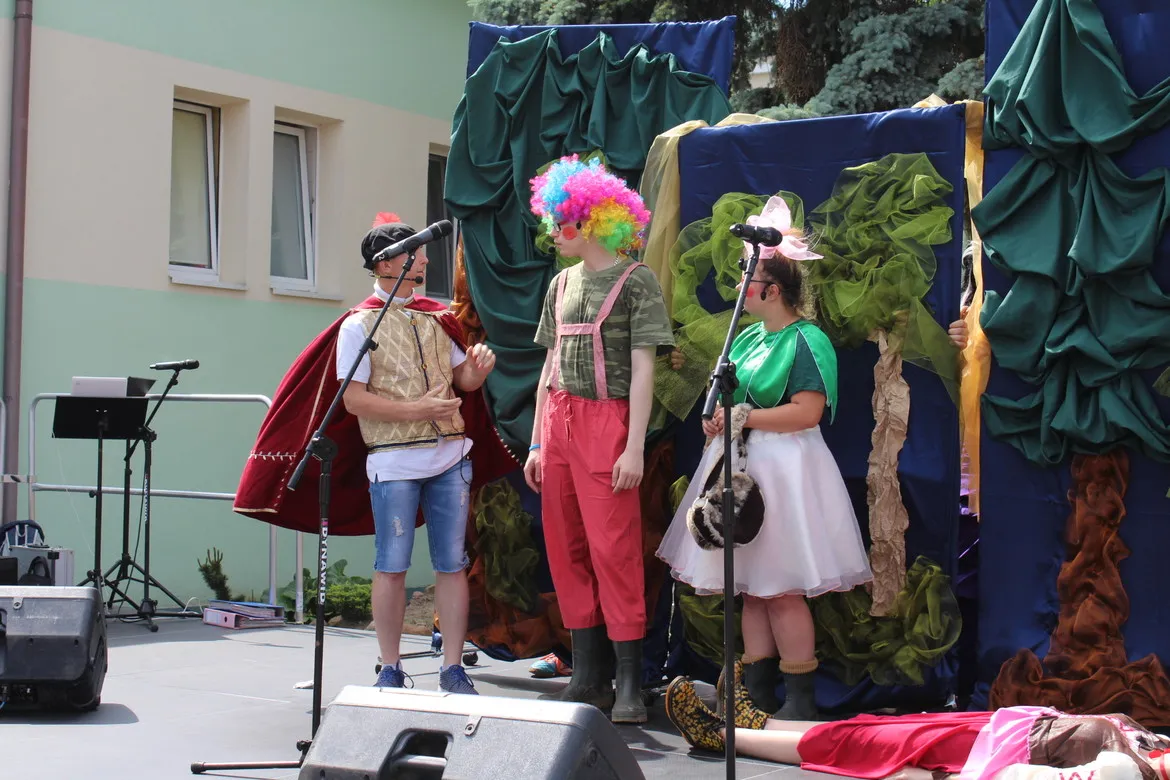 Grupa osób w kolorowych kostiumach występuje na scenie podczas Dnia Integracji i Aktywności Osób z Niepełnosprawnościami, z mikrofonami i barwną scenografią w tle.