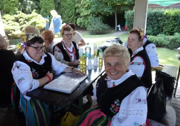 Kobiety w tradycyjnych, kolorowych strojach ludowych siedzą przy stole na świeżym powietrzu podczas plenerowego V Przeglądu Amatorskich Zespołów Artystycznych „Mikrofon Seniora”, uśmiechając się do aparatu.
