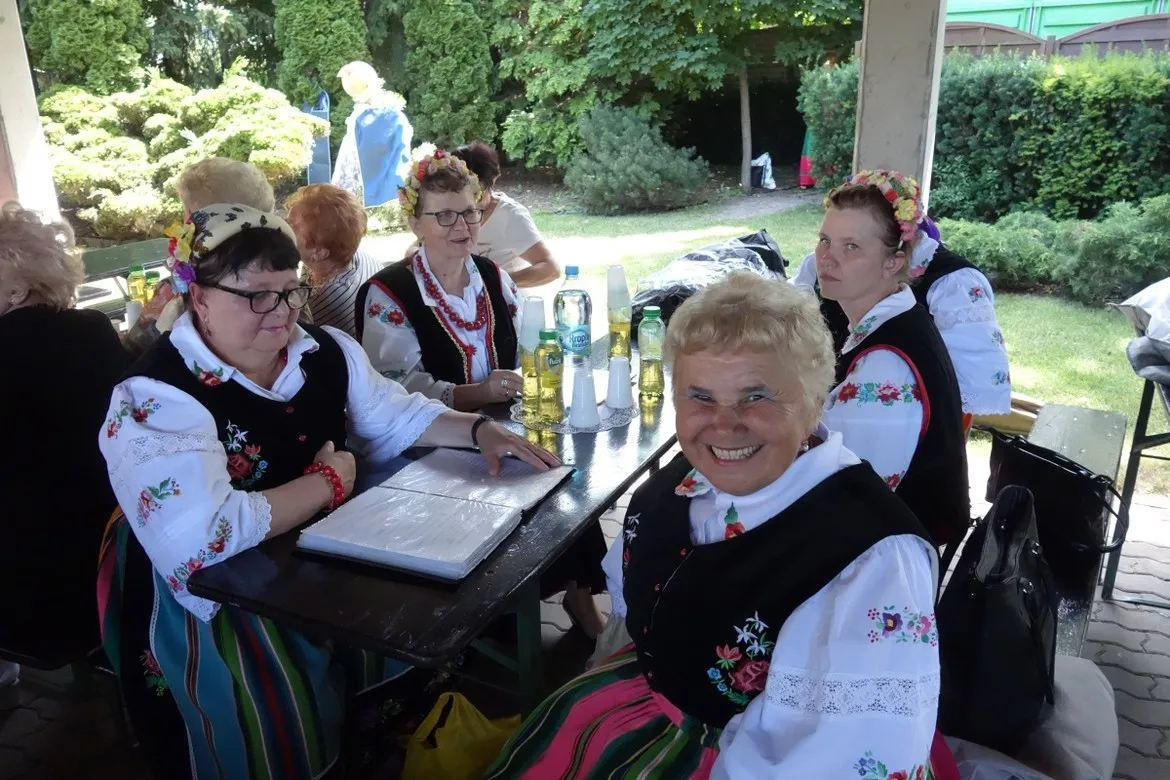 Kobiety w tradycyjnych, kolorowych strojach ludowych siedzą przy stole na świeżym powietrzu podczas plenerowego V Przeglądu Amatorskich Zespołów Artystycznych „Mikrofon Seniora”, uśmiechając się do aparatu.