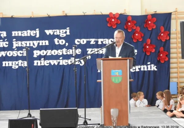 Mężczyzna w garniturze wygłasza przemówienie z podium podczas jubileuszu 100-lecia szkoły w Radzanowie, na tle niebieskiej sceny z dużymi białymi literami i czerwonymi kwiatami, a po prawej stronie siedzi grupa dziewcząt w białych bluzkach.