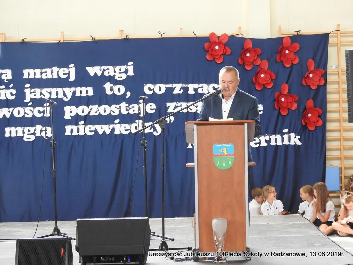 Mężczyzna w garniturze wygłasza przemówienie z podium podczas jubileuszu 100-lecia szkoły w Radzanowie, na tle niebieskiej sceny z dużymi białymi literami i czerwonymi kwiatami, a po prawej stronie siedzi grupa dziewcząt w białych bluzkach.