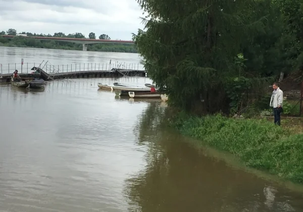 Mężczyzna stoi na brzegu rzeki w Powiecie Płockim, obok uszkodzonego pomostu i kilku łodzi, a w tle widać most.