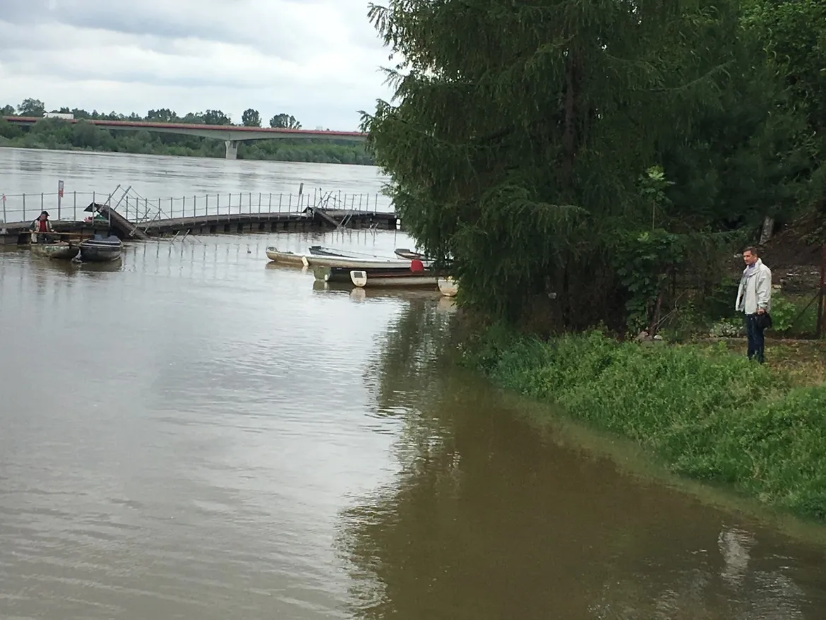 Mężczyzna stoi na brzegu rzeki w Powiecie Płockim, obok uszkodzonego pomostu i kilku łodzi, a w tle widać most.