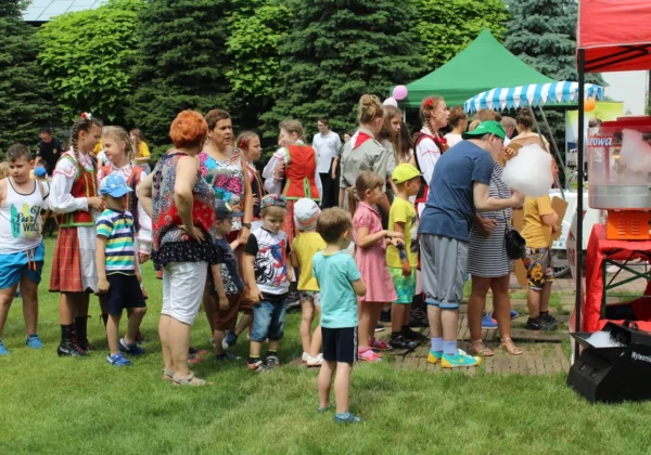 Grupa dorosłych i dzieci na plenerowym festynie, część w ludowych strojach, stoi w kolejce do stoiska z watą cukrową.