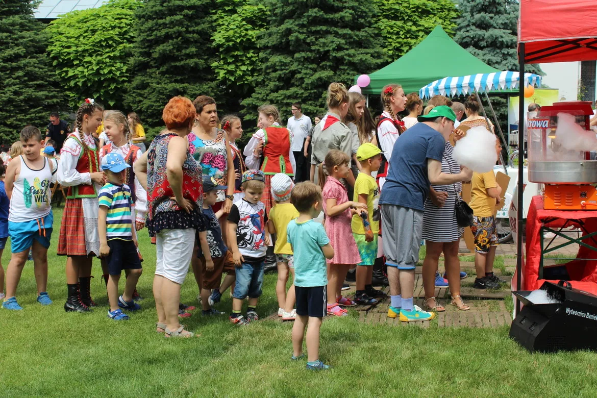 Grupa dorosłych i dzieci na plenerowym festynie, część w ludowych strojach, stoi w kolejce do stoiska z watą cukrową.