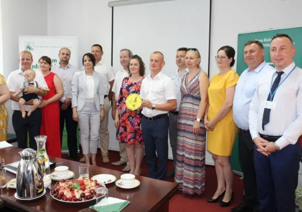 Grupa osób stoi na scenie w sali konferencyjnej podczas rozstrzygnięcia konkursu Bezpieczne Gospodarstwo Rolne 2019, a mężczyzna w białej koszuli trzyma żółtą okrągłą nagrodę.