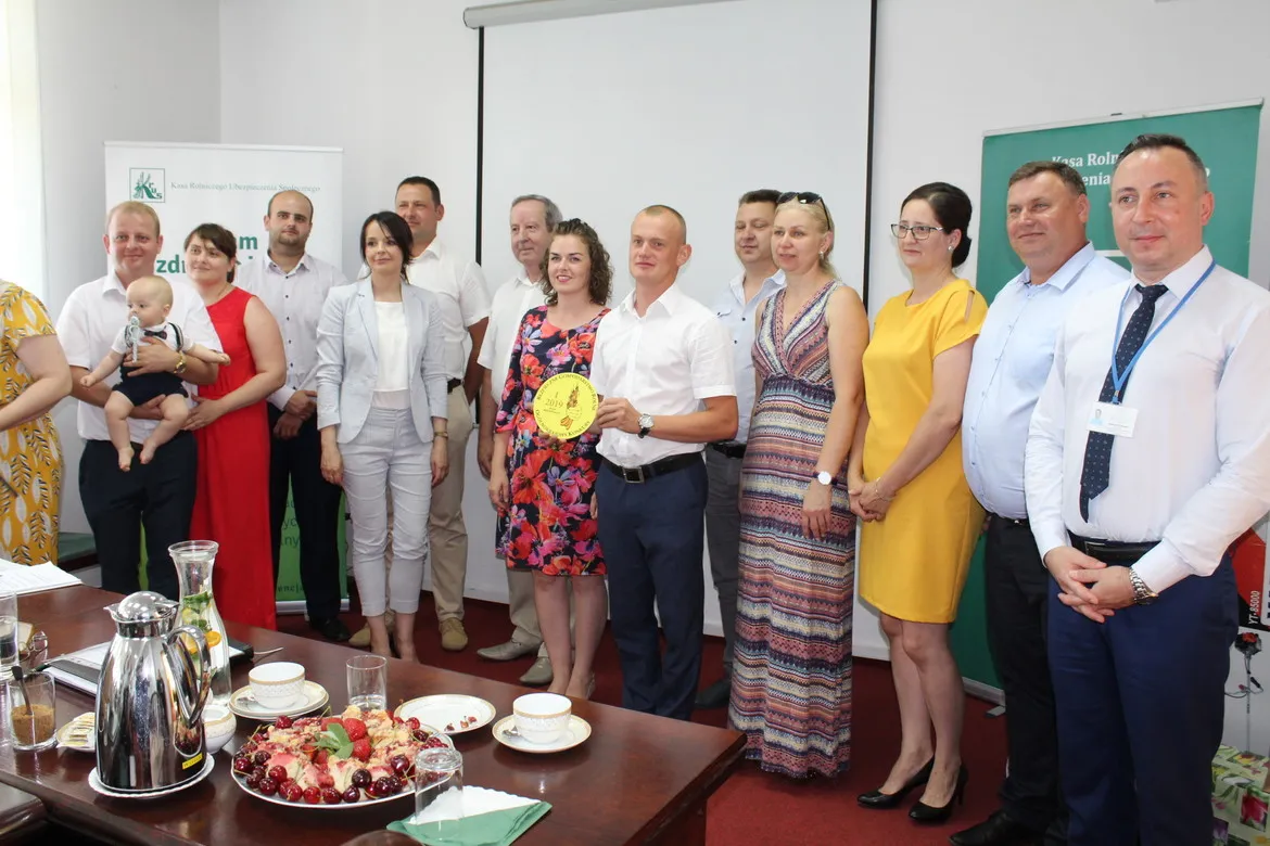 Grupa osób stoi na scenie w sali konferencyjnej podczas rozstrzygnięcia konkursu Bezpieczne Gospodarstwo Rolne 2019, a mężczyzna w białej koszuli trzyma żółtą okrągłą nagrodę.