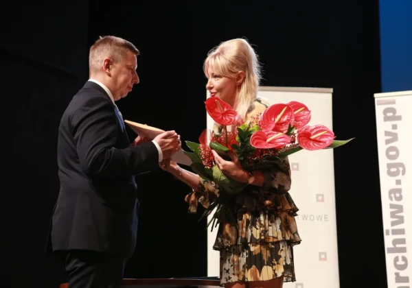 Na scenie podczas obchodów Stulecia Archiwum Państwowego w Płocku mężczyzna w garniturze wręcza nagrodę kobiecie, która trzyma duży bukiet czerwonych kwiatów anthurium.