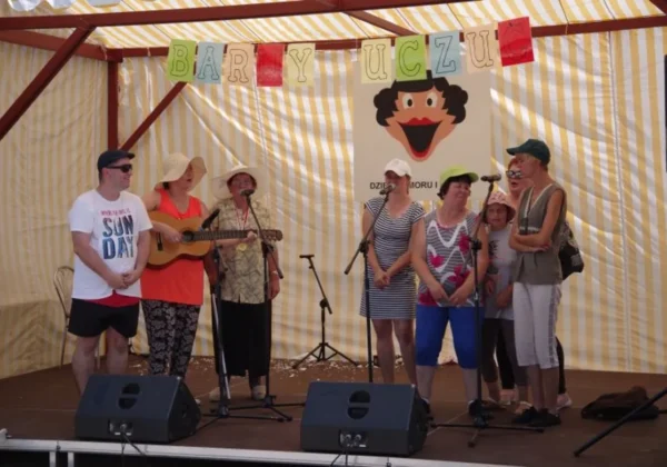 Grupa ludzi na scenie w żółto‑białym namiocie, śpiewająca i grająca na gitarze podczas XXVI Dnia Humoru i Satyry w Zakrzewie, obok mikrofonów i plakatu z wizerunkiem uśmiechniętego klauna.