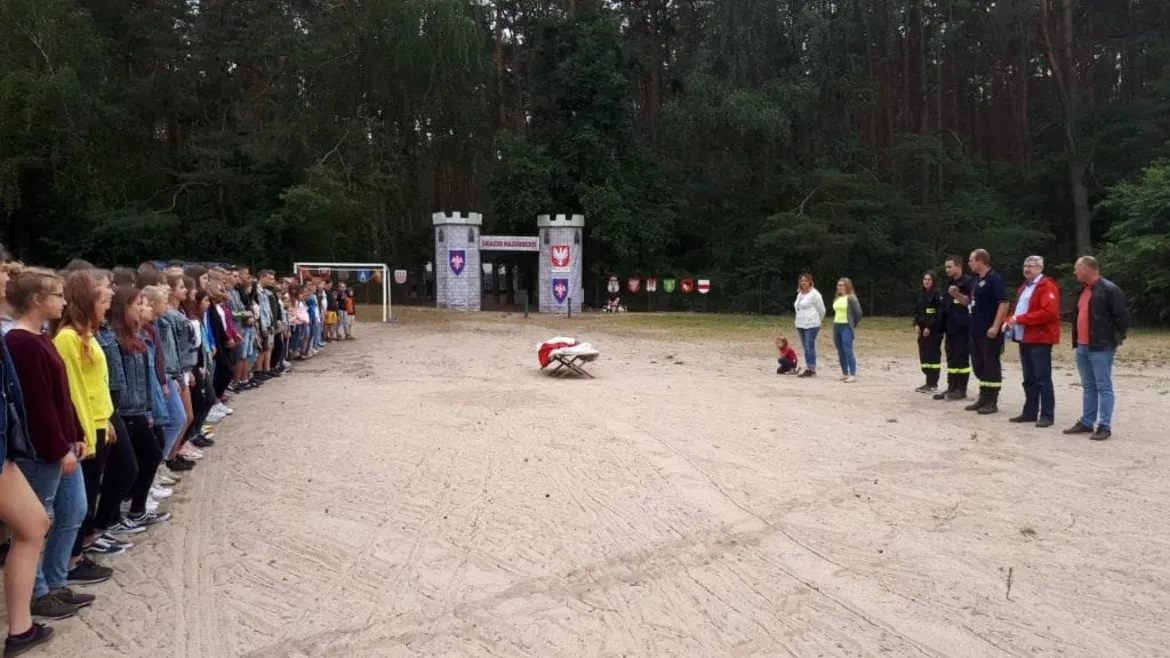 Podczas Obózu Szkoleniowo-Wypoczynkowego MDP 2019 grupa młodzieży stoi w dwóch rzędach po obu stronach placu, a na środku znajduje się osoba na noszach symulująca akcję ratunkową.