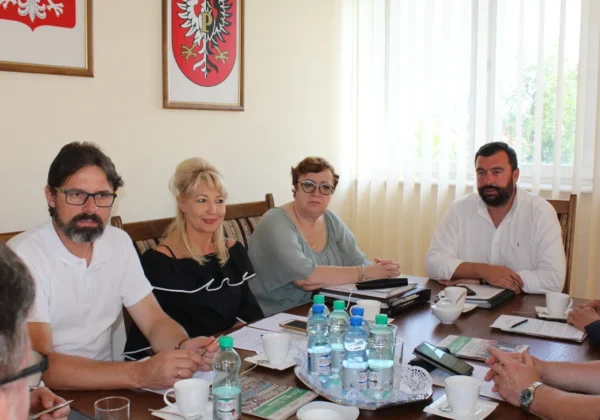 Czterech członków Zarządu Powiatu Płockiego siedzi przy stole podczas spotkania, mają przed sobą dokumenty i napoje, a na ścianie widać herby powiatu.