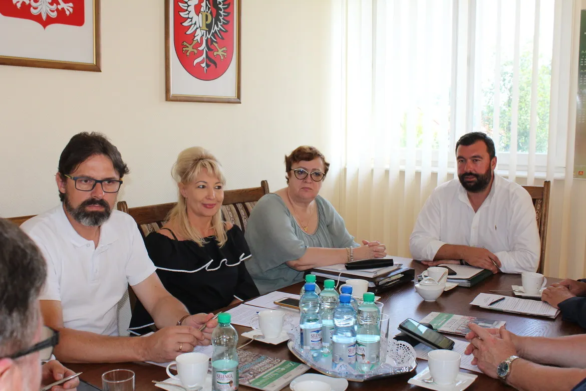 Czterech członków Zarządu Powiatu Płockiego siedzi przy stole podczas spotkania, mają przed sobą dokumenty i napoje, a na ścianie widać herby powiatu.