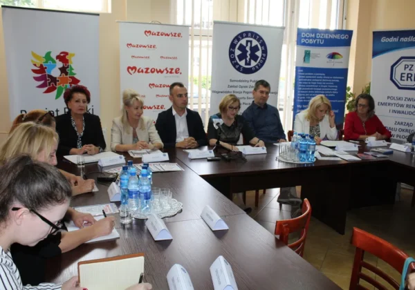 Grupa osób siedzi przy długim stole podczas spotkania na temat „Świadomy i bezpieczny senior – inwestycją w życie i zdrowie”, z bannerami organizacji w tle.