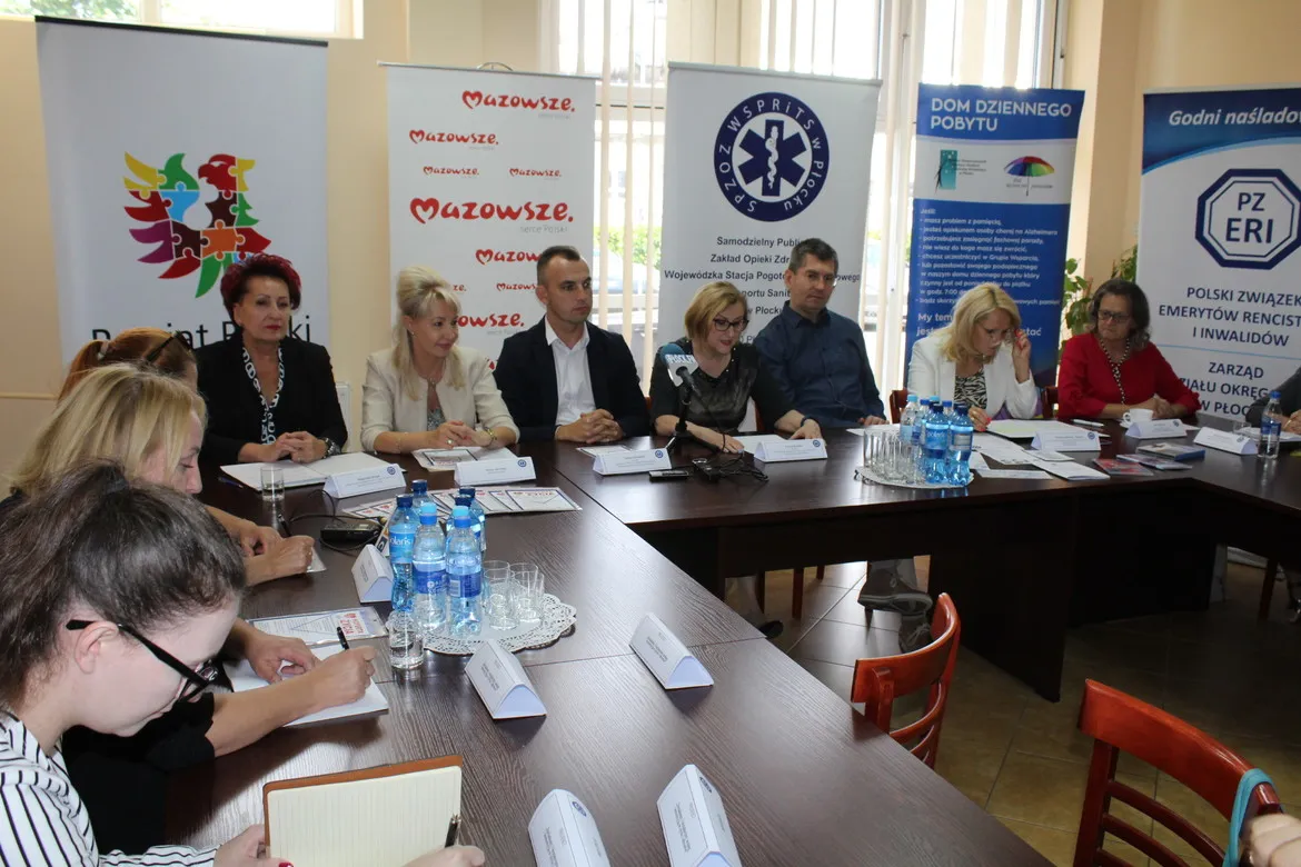 Grupa osób siedzi przy długim stole podczas spotkania na temat „Świadomy i bezpieczny senior – inwestycją w życie i zdrowie”, z bannerami organizacji w tle.