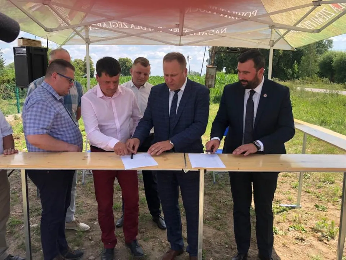 Grupa osób podpisuje dokumenty na drewnianym stole podczas ceremonii rozpoczęcia remontu ostatniego odcinka drogi Bulkowo – Worowice – granica powiatu, pod namiotem w plenerze.