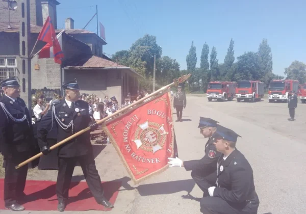Uroczystość strażacka w powiecie płockim — strażacy w ceremonialnych mundurach prezentują sztandar przed grupą widzów i stojącymi wozami gaśniczymi.