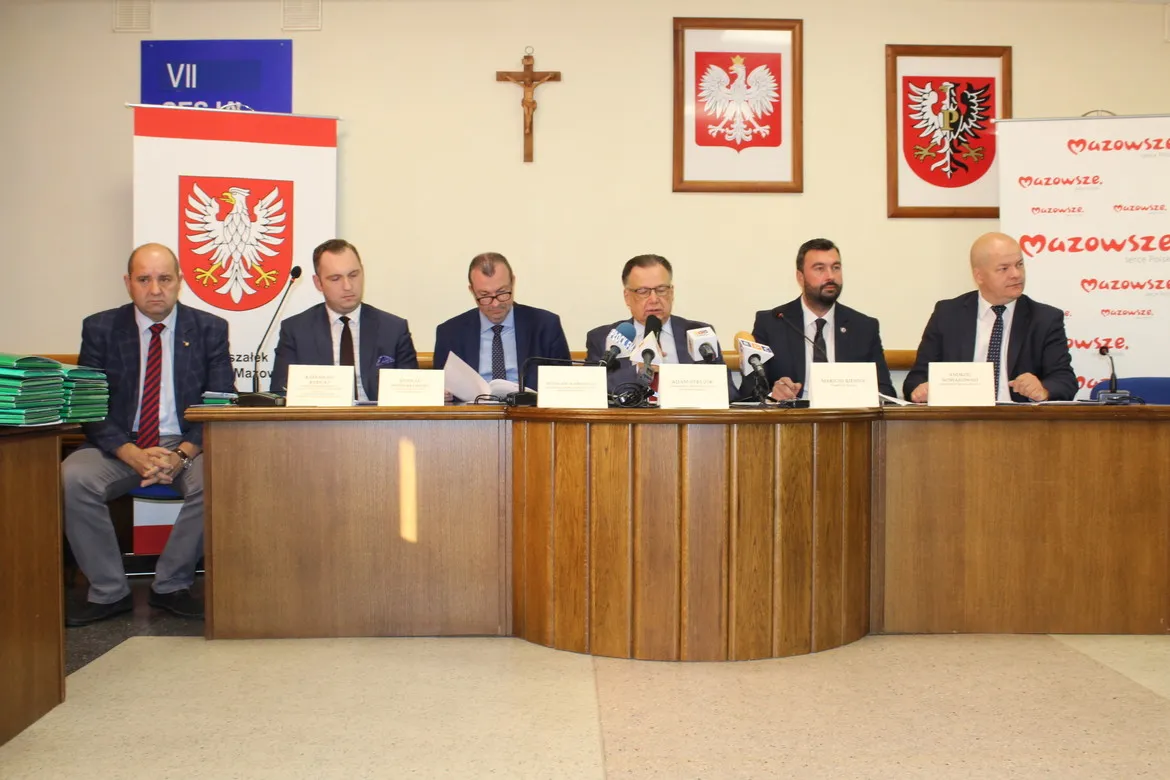 Grupa urzędników w garniturach siedzi za drewnianym, zakrzywionym stołem podczas konferencji prasowej o dofinansowaniu Powiatu Płockiego z budżetu Mazowsza (ponad 2 mln zł), z mikrofonami i flagami w tle.