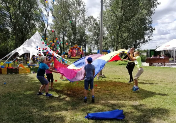 Dzieci i dorośli bawią się dużą, kolorową parachutą podczas pikniku integracyjnego w parku, w tle znajdują się namioty, balony i atrakcje.