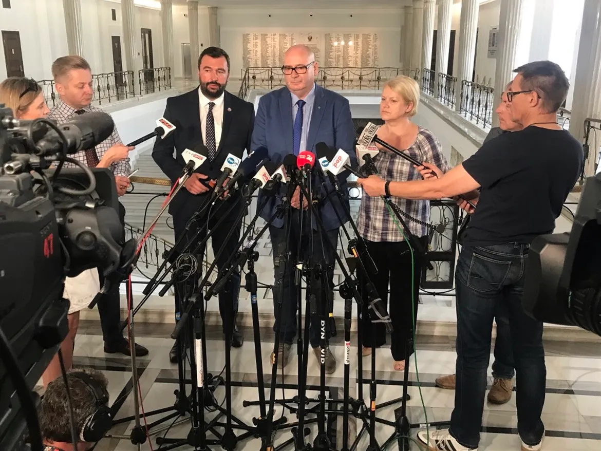 Grupa urzędników udziela konferencji prasowej dotyczącej AFS, otoczona przez reporterów i liczne mikrofony.